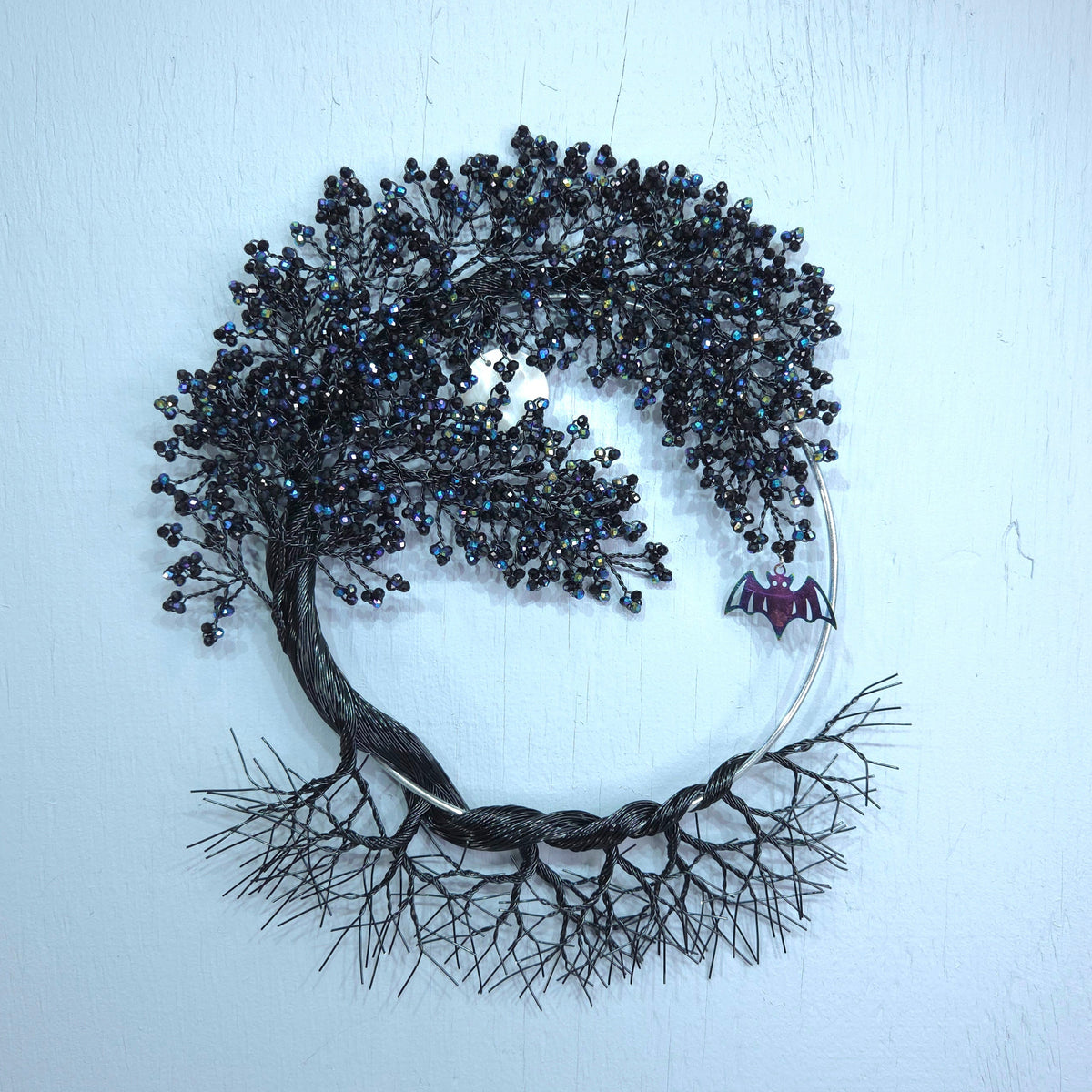 A circular black wire wreath wall hanging featuring a stylized tree with dark brown and black branches, black seed-like beads, and a small purple bat decoration on the right side, displayed against a light blue wall.