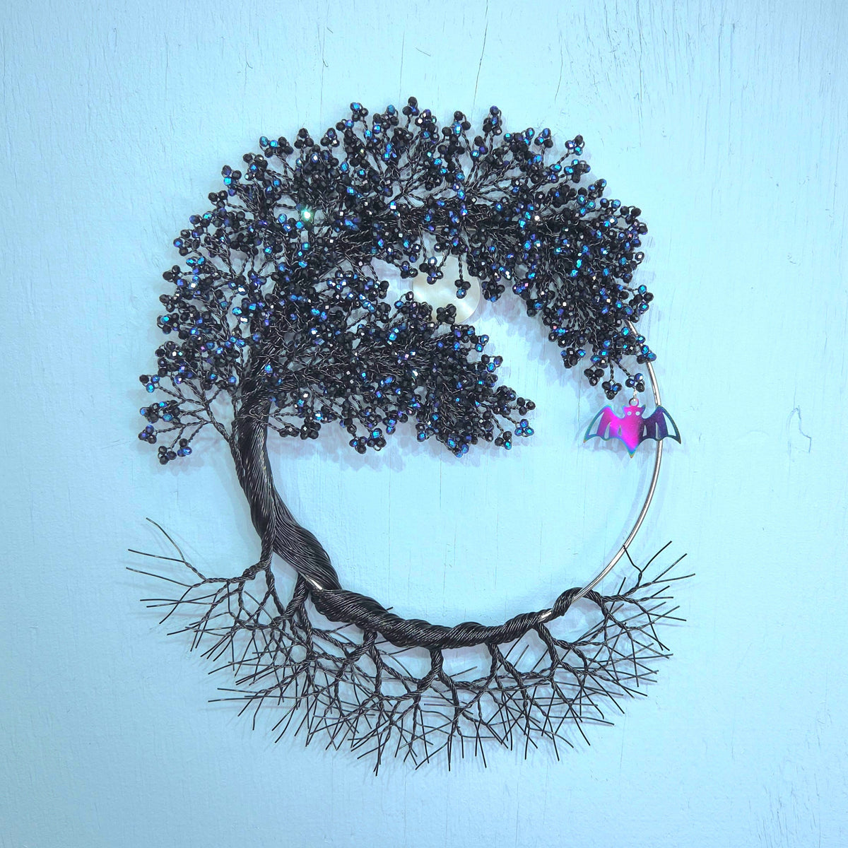 A circular metal wall art piece shaped like a tree with dark metallic branches and leaves, accented by small blue iridescent beads and a small pink bat on the right side, mounted against a light blue wall.