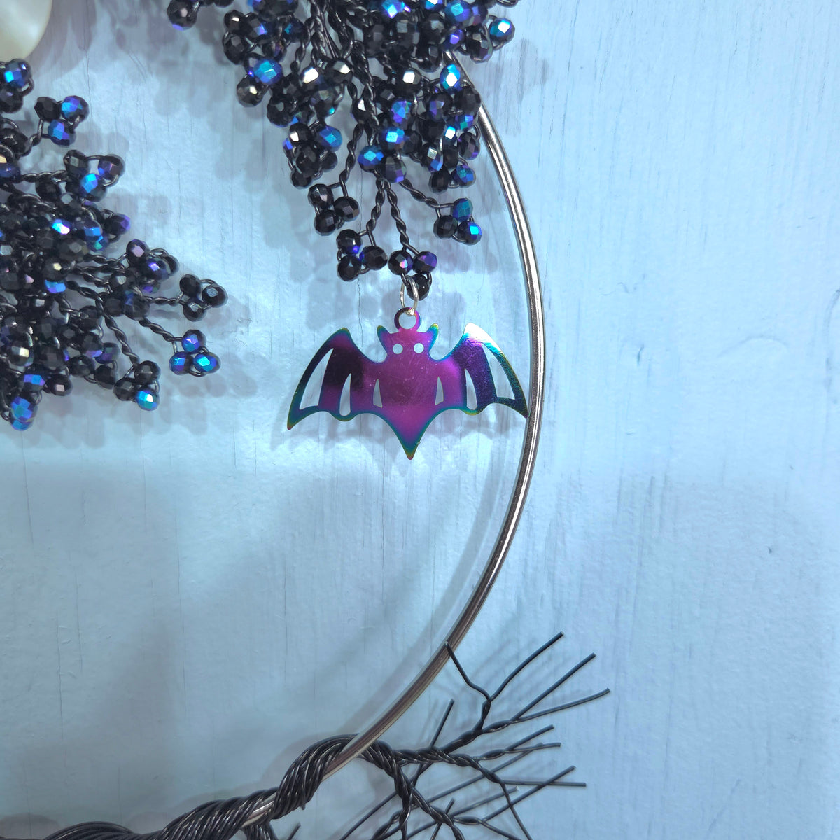A close-up of a bat-shaped pendant with an iridescent purple and pink finish, hanging from a thin metal hoop. The hoop is decorated with black and blue beaded branches against a pale blue background.