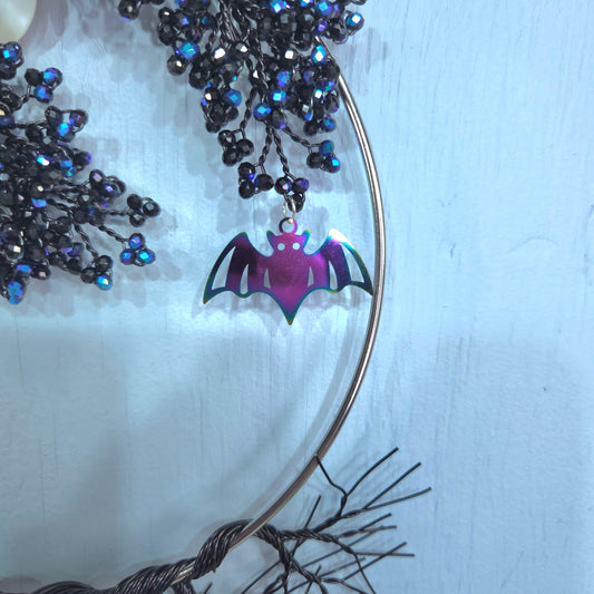 A close-up of a bat-shaped pendant with an iridescent purple and pink finish, hanging from a thin metal hoop. The hoop is decorated with black and blue beaded branches against a pale blue background.