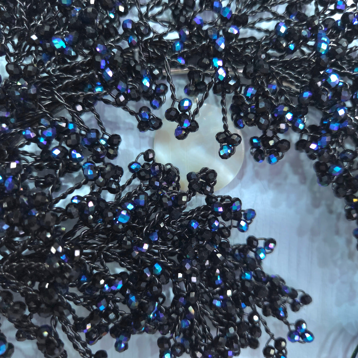 A close-up of a beaded garland featuring black seed beads with iridescent blue and purple AB finish, draped over a white circular display stand.