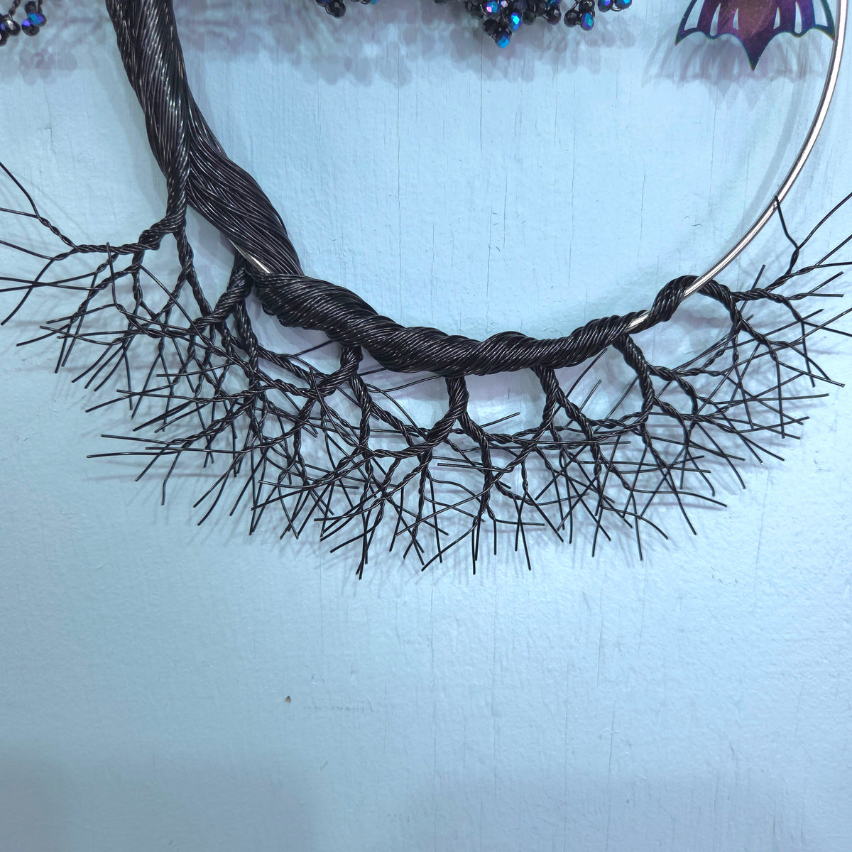 A close-up of a dark brown, branch-like decorative wreath or wall hanging made from twisted wire and many thin spindly branches, attached to a metal hoop against a pale blue wall.