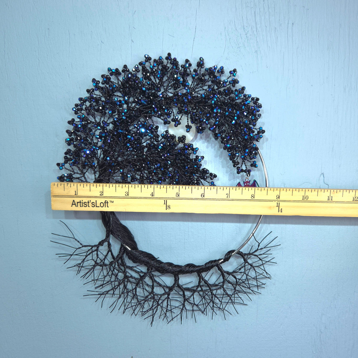 A black beaded tree-of-life wall hanging with a braided trunk and root system, shown next to a wooden ruler on a light blue wall. The ruler shows the brand 'Artist'sLoft'.