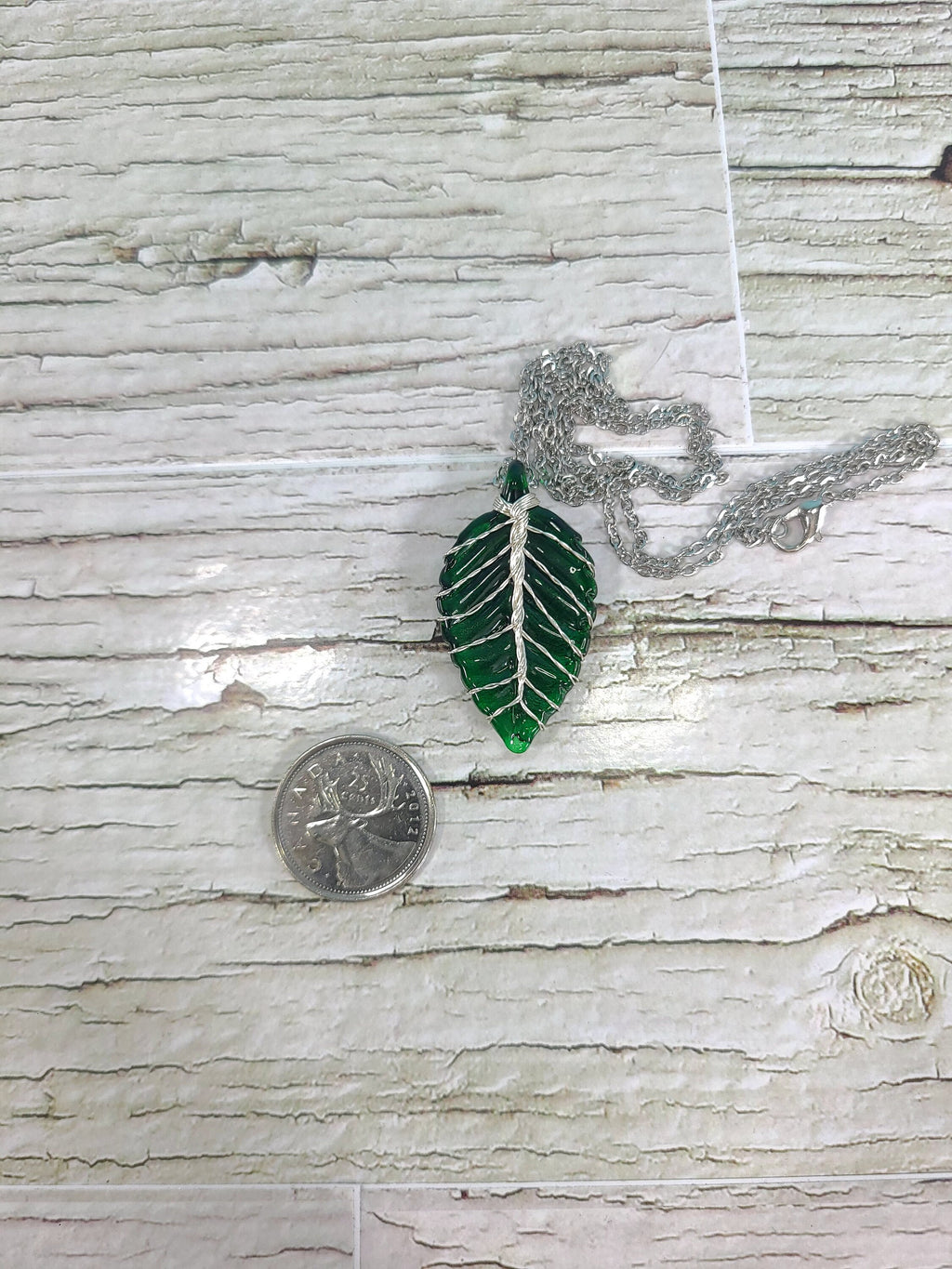 Green leaf-shaped pendant on a chain next to a quarter for scale on a wooden surface
