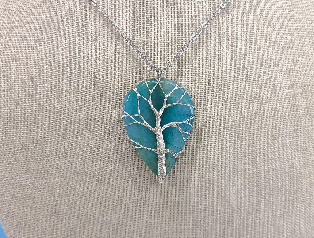 Teal tree-shaped pendant on a silver chain against a beige fabric background