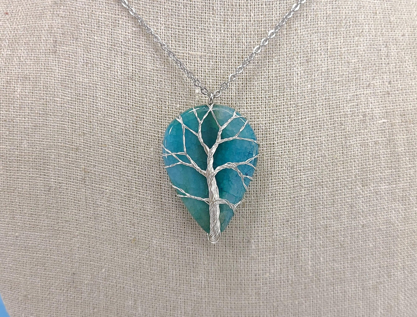 Teal tree-shaped pendant on a silver chain against a beige fabric background