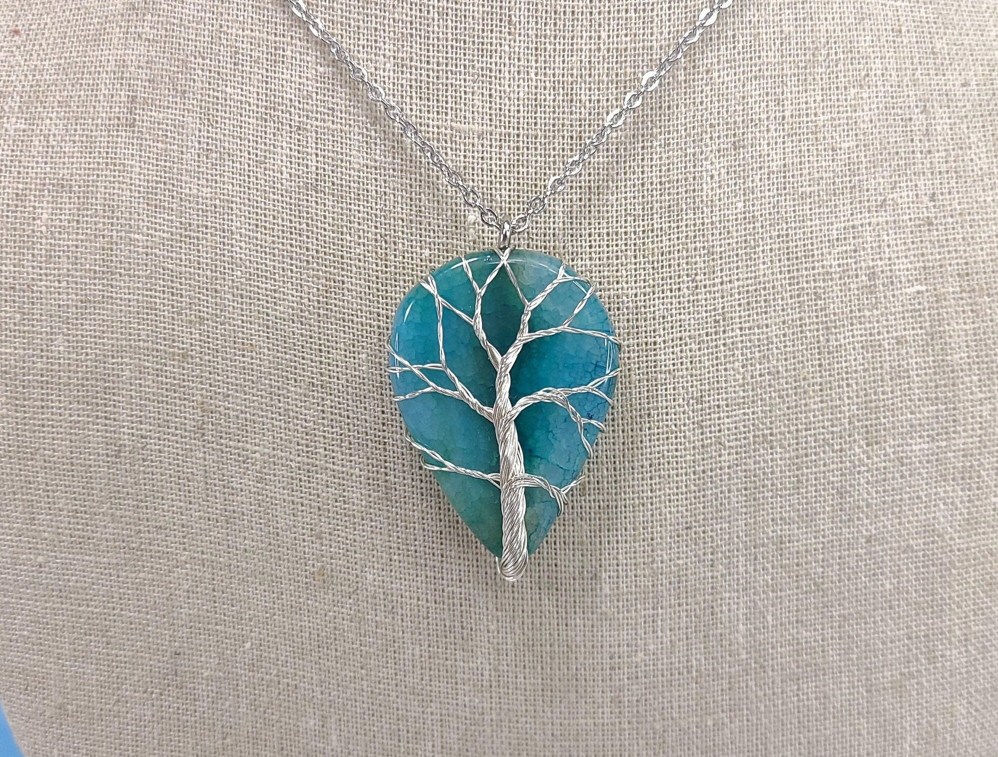 Teal tree-shaped pendant on a silver chain against a beige fabric background