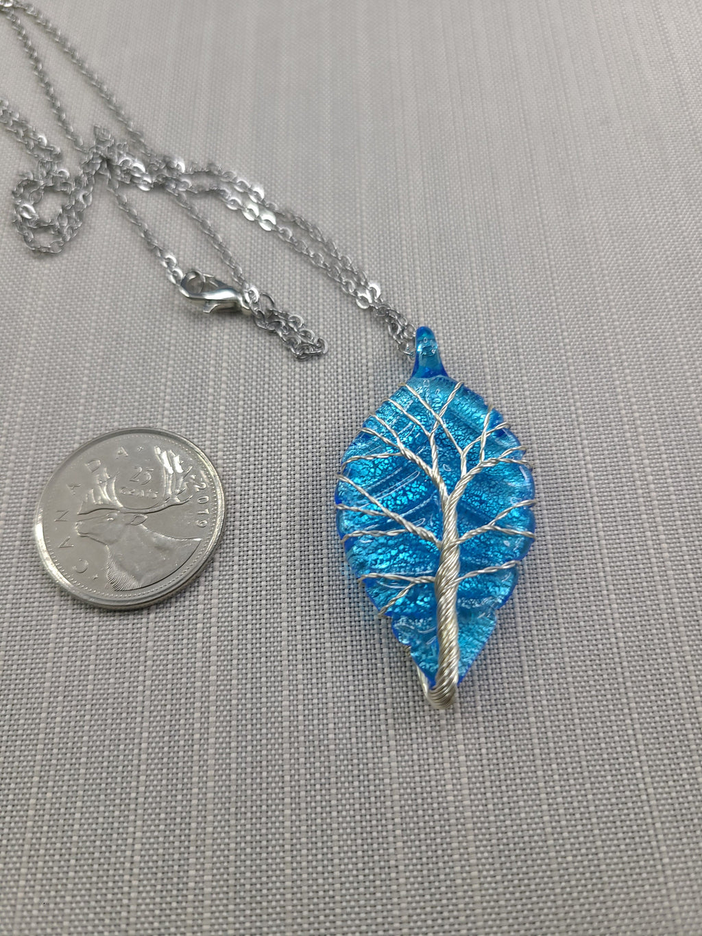 Blue leaf-shaped pendant with silver tree design on a gray background