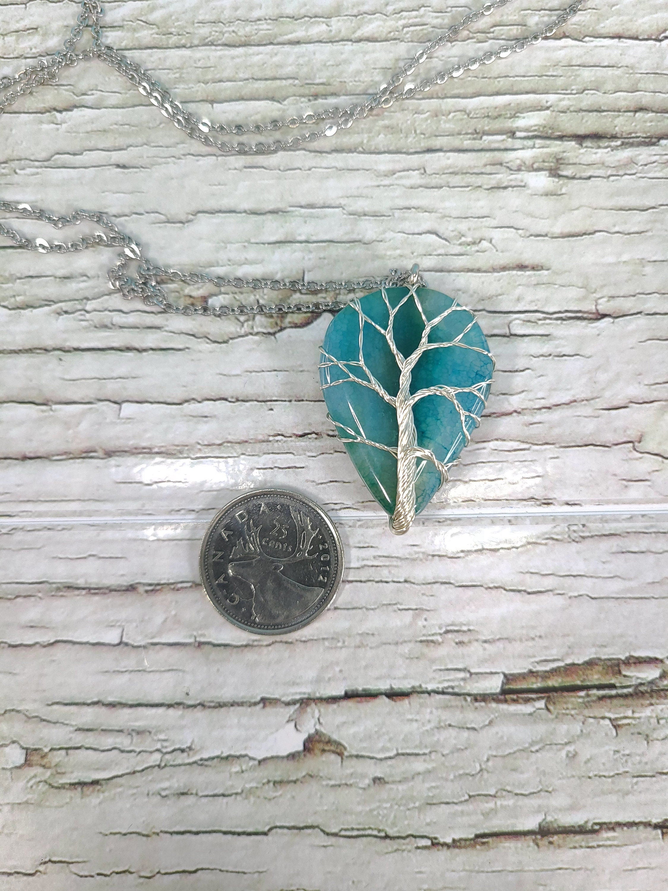 Teal leaf-shaped pendant with silver branch design on a chain, next to a quarter for scale, on a wooden surface.