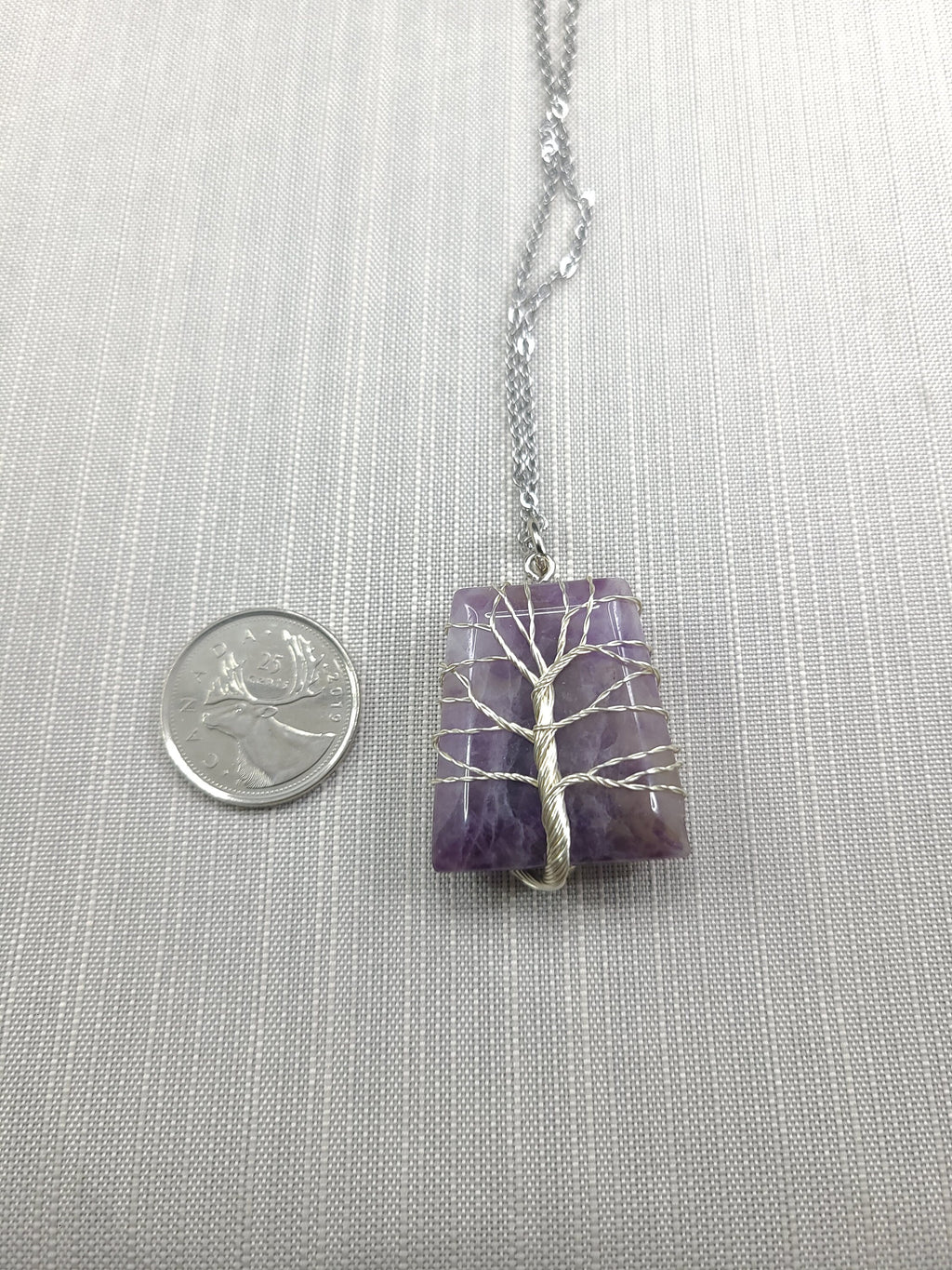 Necklace with a purple pendant featuring a white tree design, next to a quarter for scale, on a gray fabric background.
