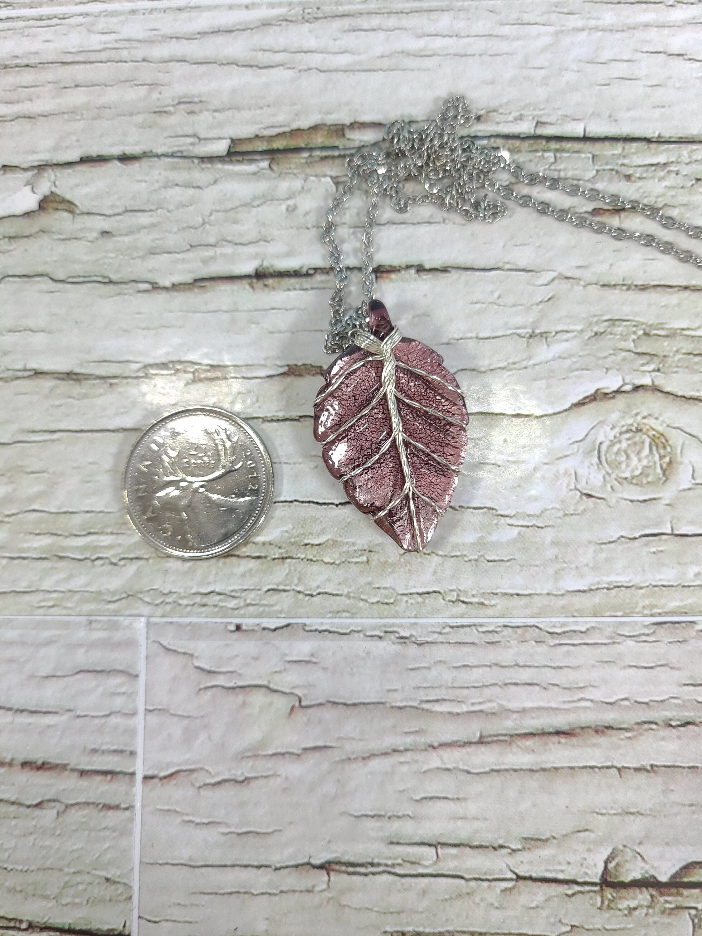 Pink leaf-shaped pendant on a chain next to a quarter for scale, on a wooden surface.