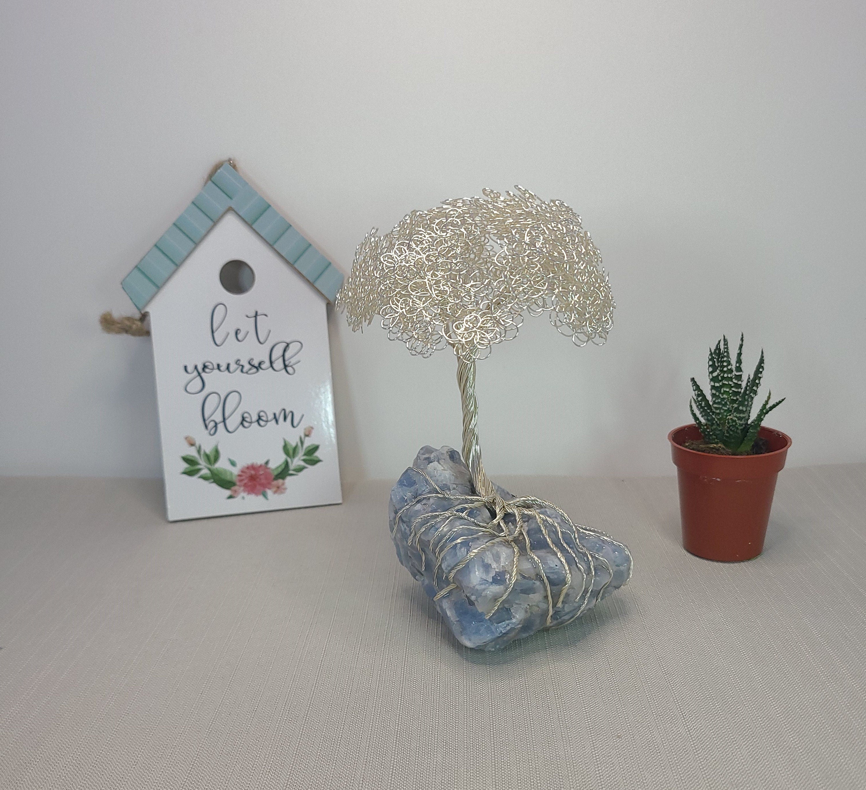 Decorative items including a small house-shaped sign, wire tree sculpture, and potted plant on a neutral background.
