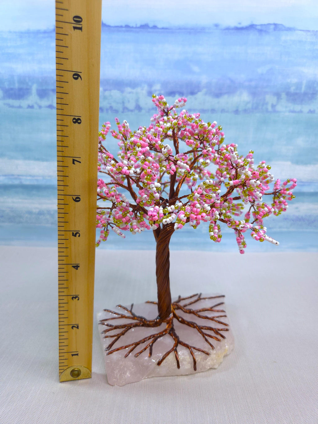 Small decorative tree with pink blossoms on a clear base, next to a ruler for scale, against a blue and white background.