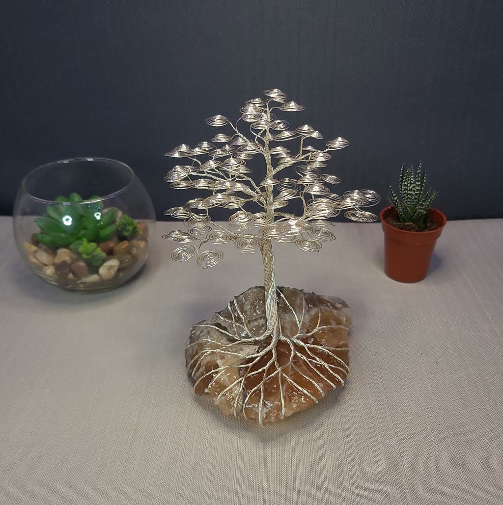 Decorative wire tree on a stone base with two potted plants in the background.