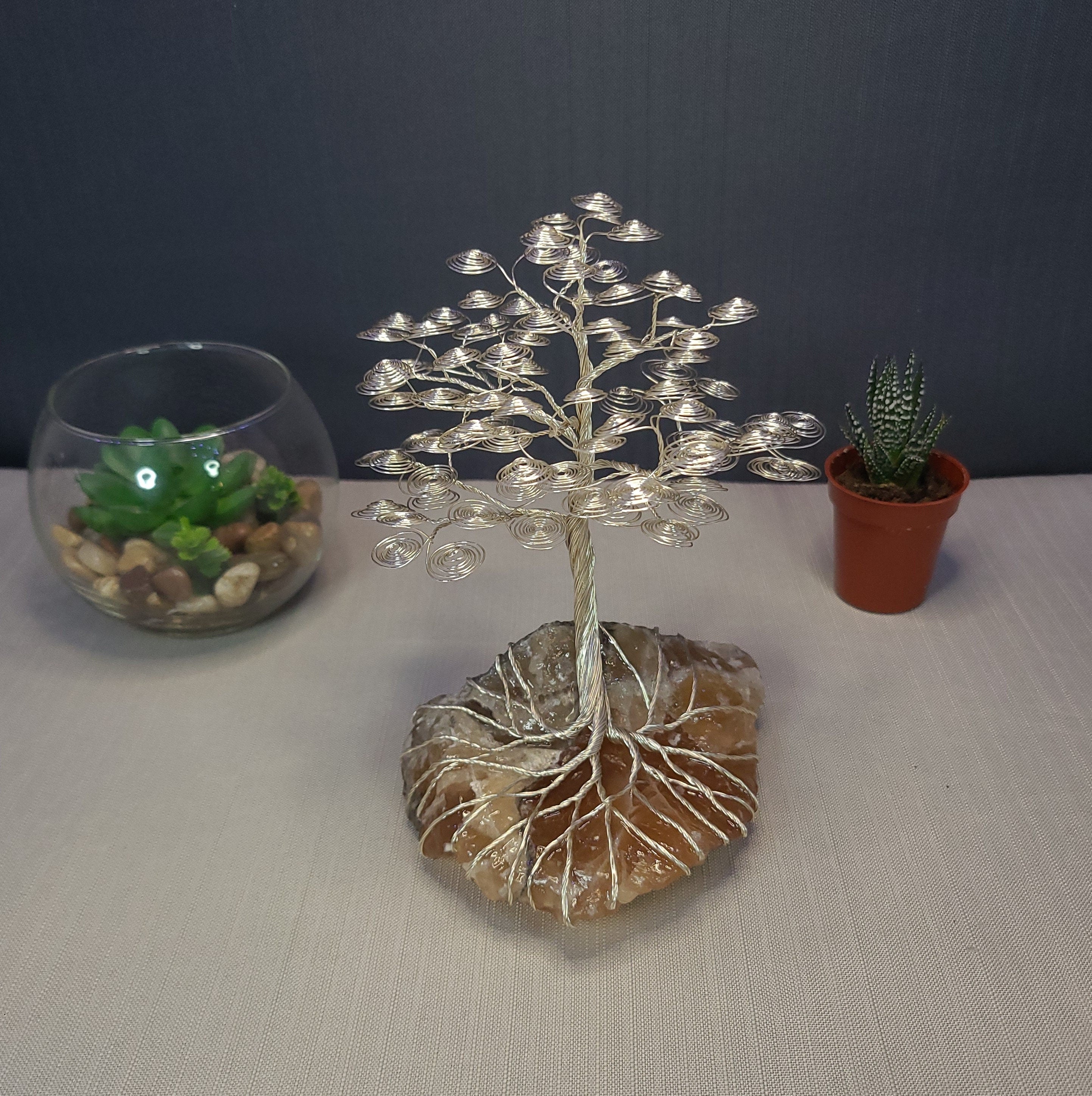 Decorative wire tree on a stone base with two potted plants in the background.