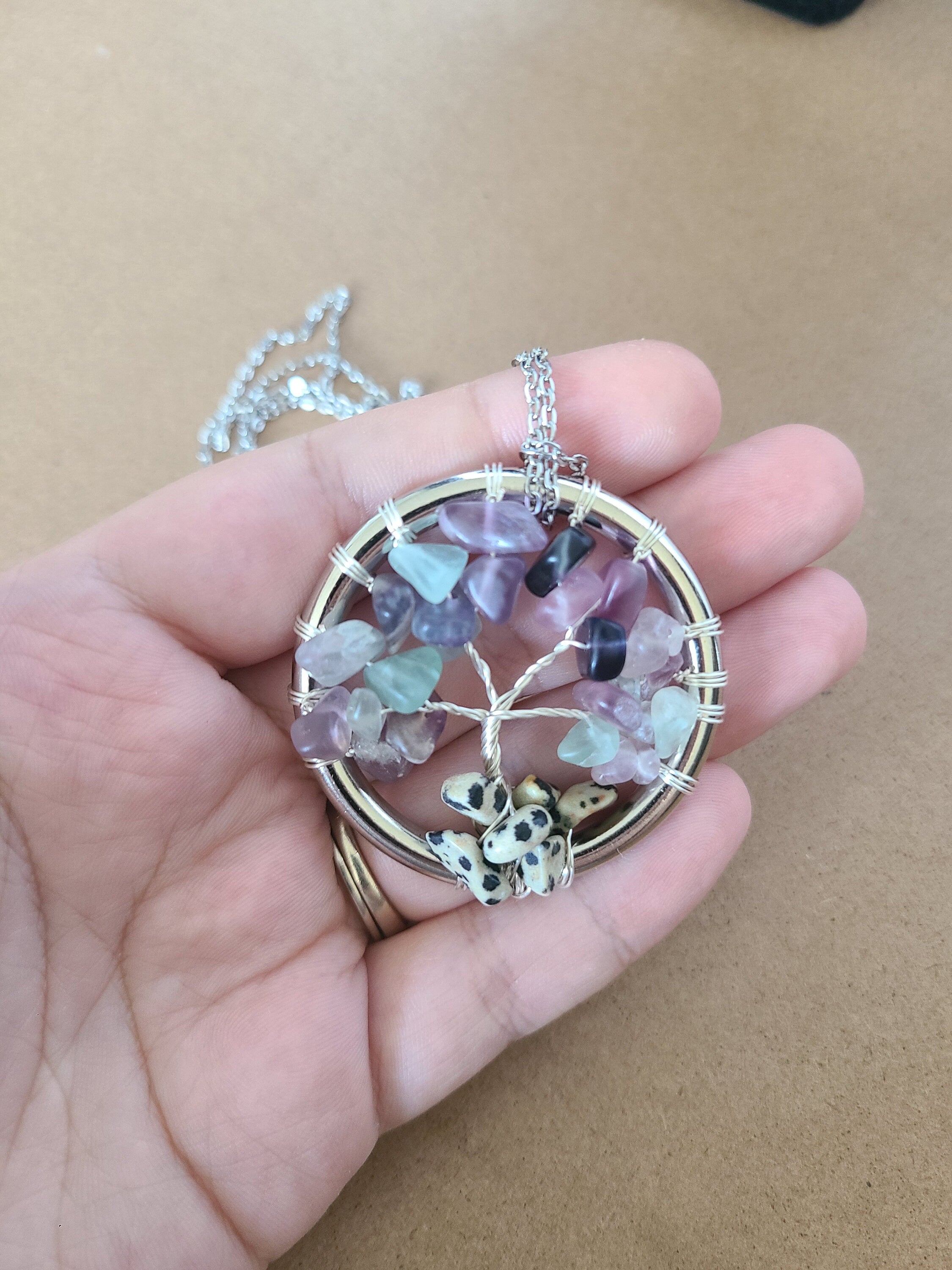 Hand holding a tree of life pendant with colorful stones against a brown background