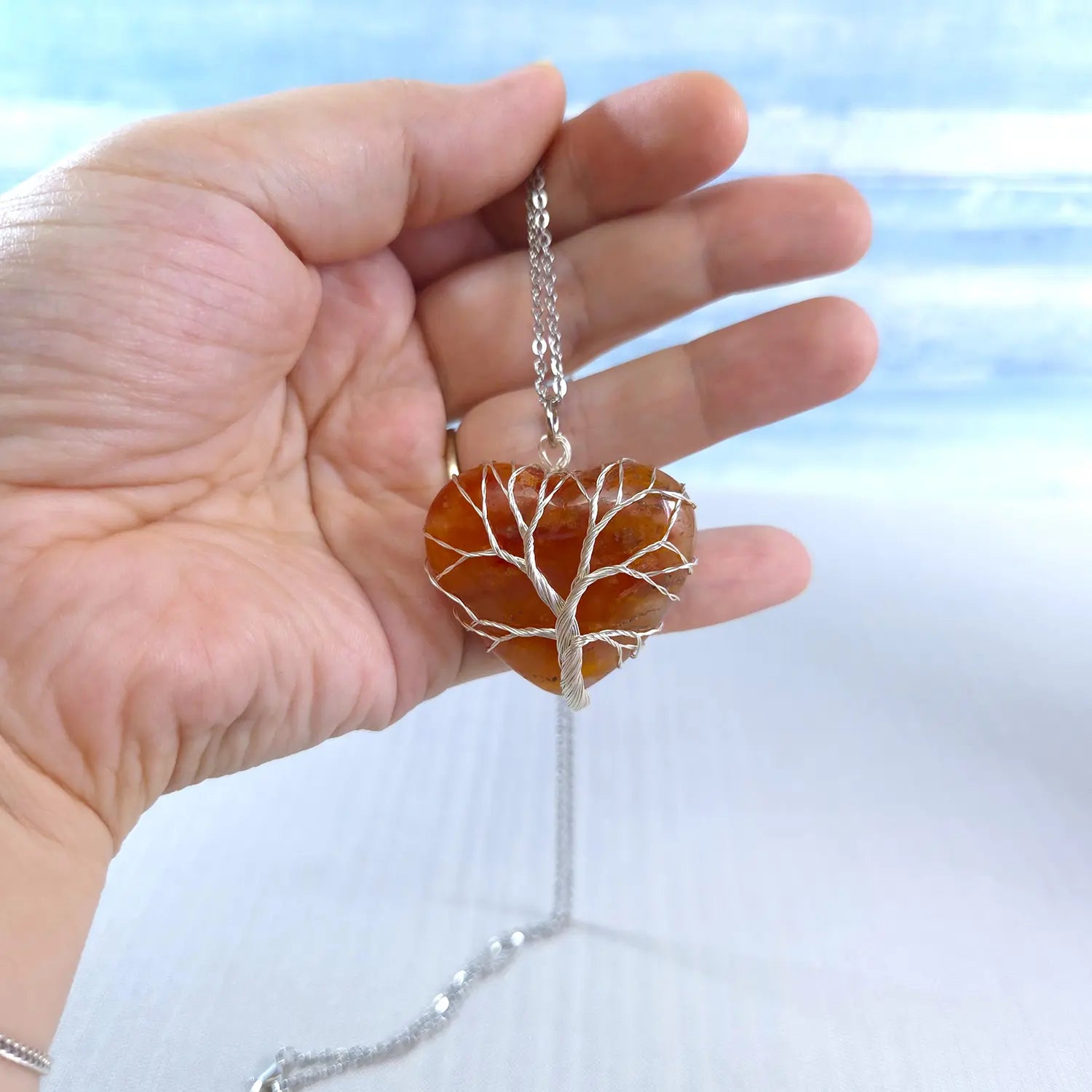 Hand holding a necklace with a heart-shaped tree pendant against a light background