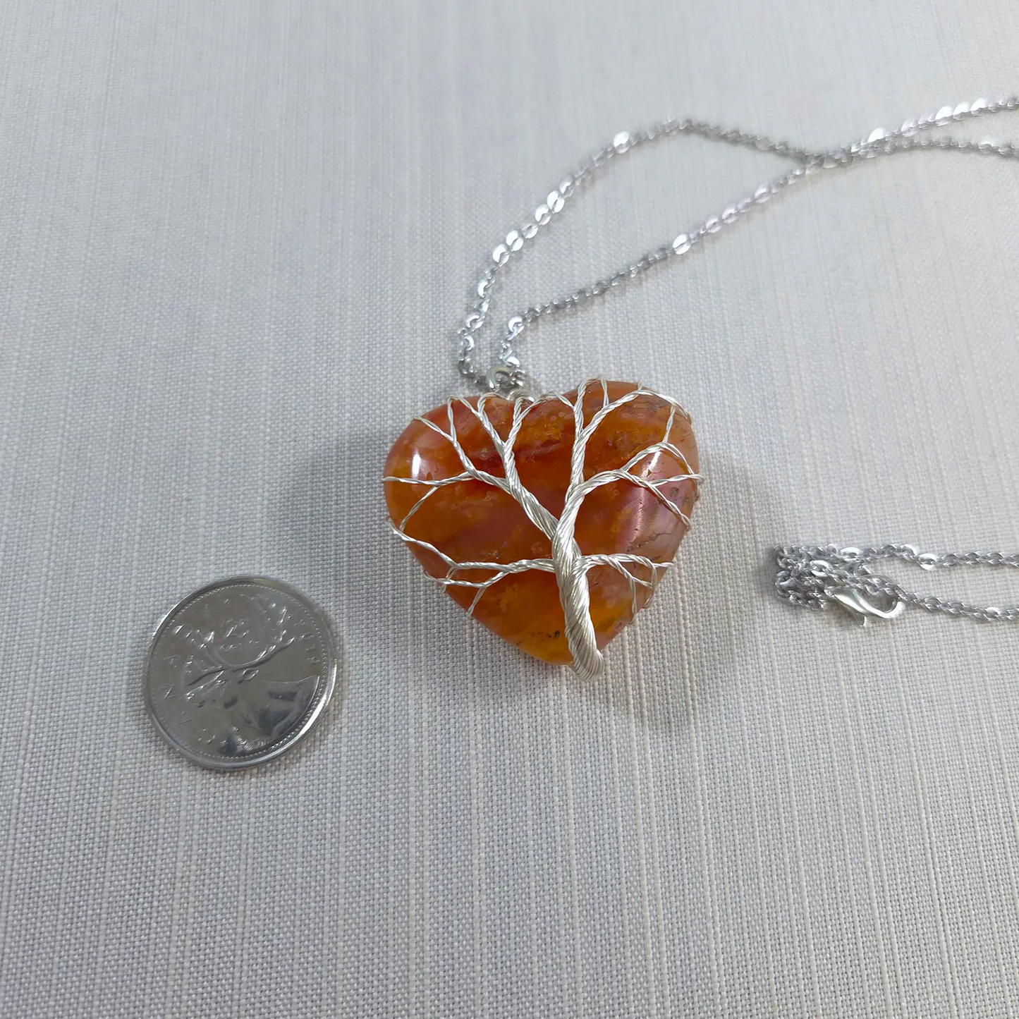 Heart-shaped amber pendant with silver tree design on a chain, next to a coin for scale.