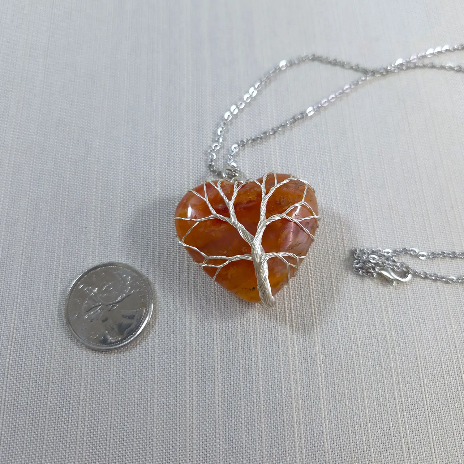 Heart-shaped amber pendant with silver tree design on a chain, next to a coin for scale.