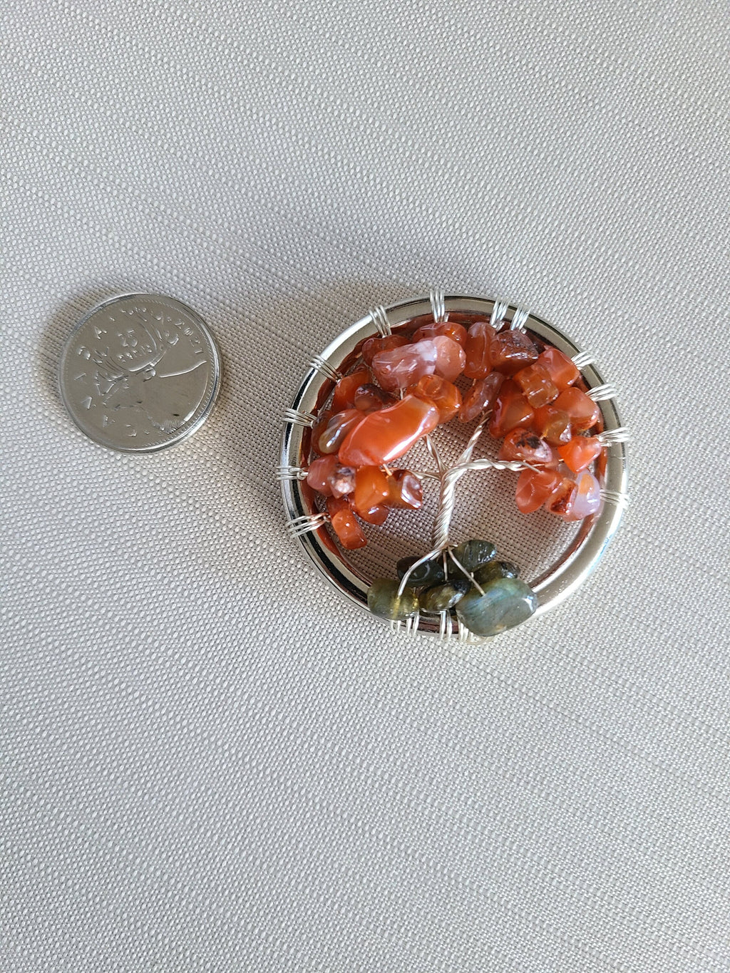 Small tree of life jewelry piece with orange and green stones next to a coin on a gray background