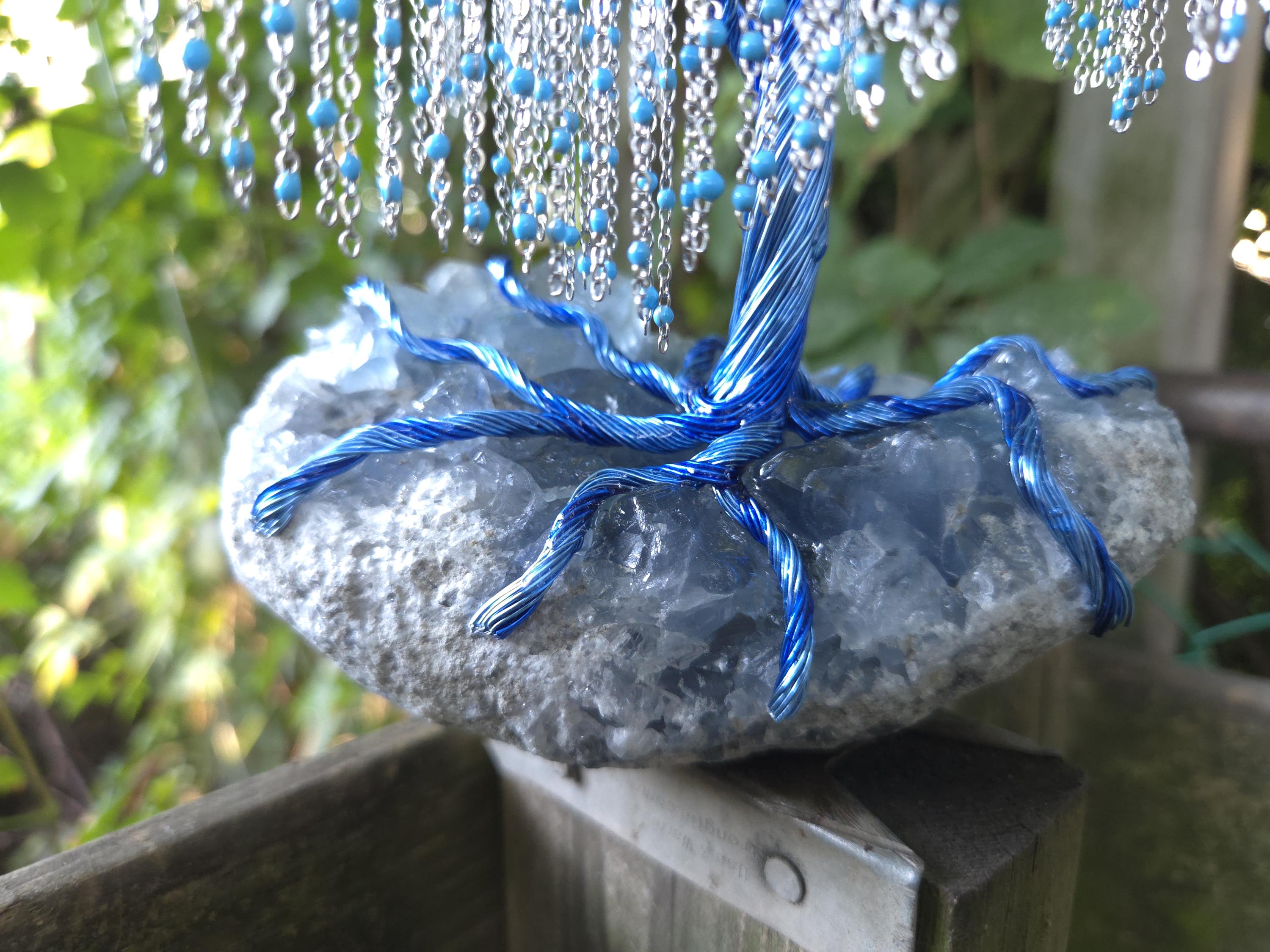 Decorative stone with blue beads and ribbons on a wooden surface with greenery in the background