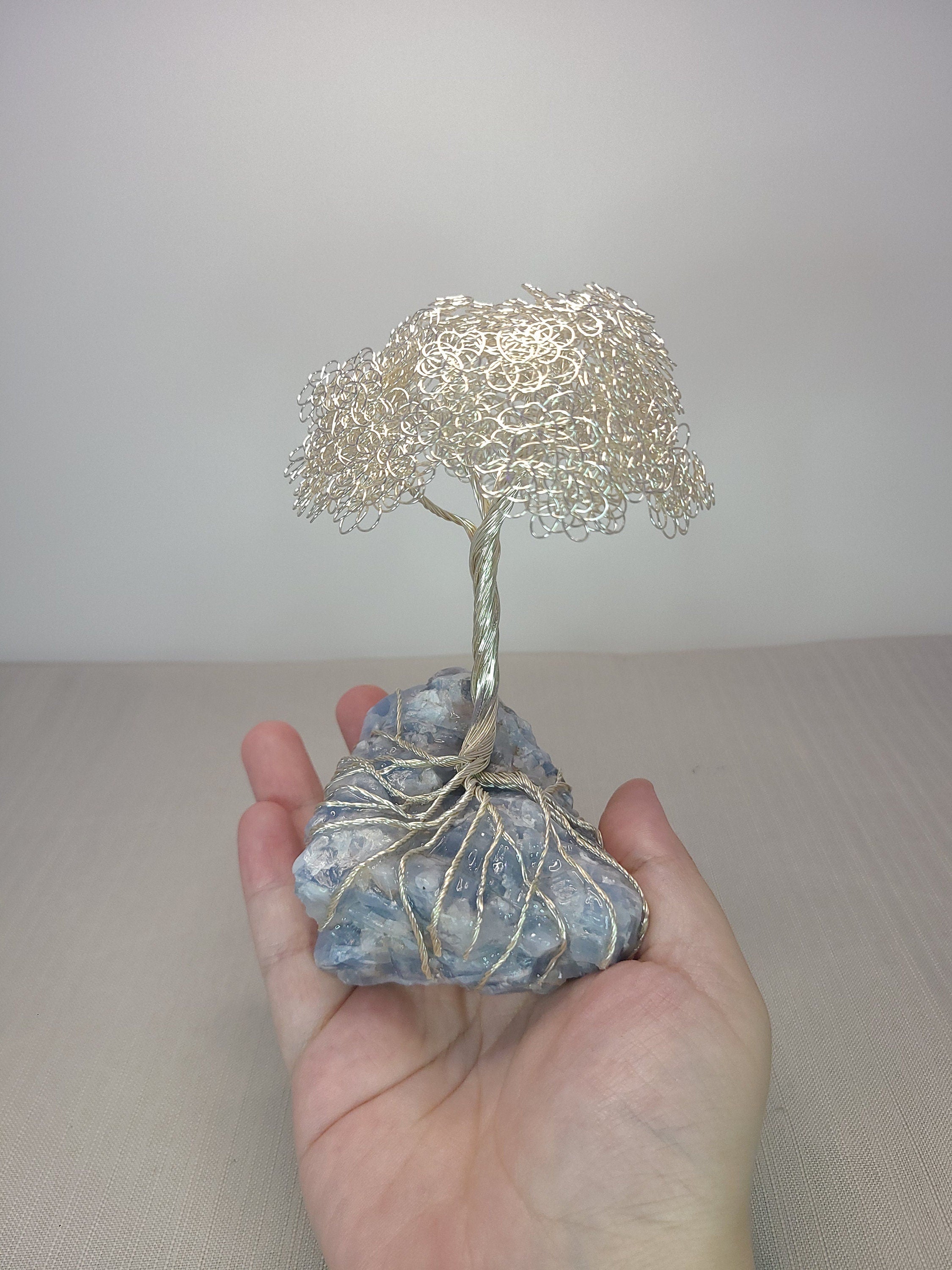 Hand holding a small wire tree sculpture on a rock against a plain background