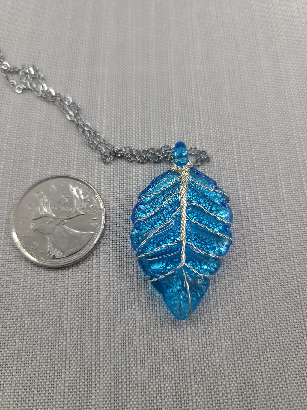 Blue leaf-shaped pendant on a chain with a quarter for scale on a gray background