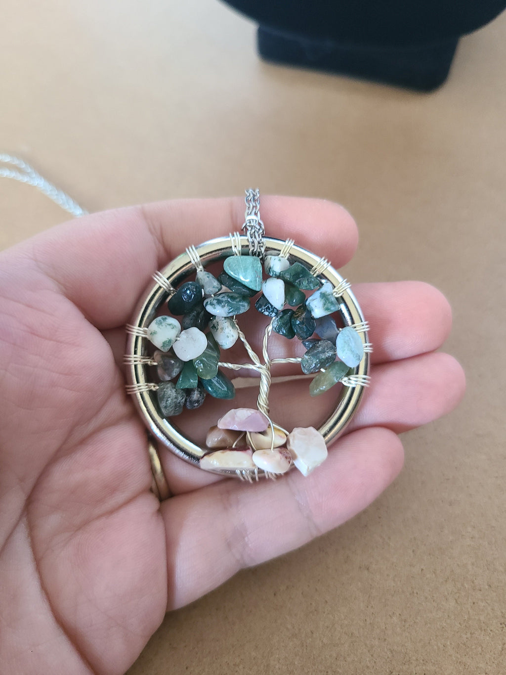 Hand holding a tree of life pendant with multicolored stones against a neutral background