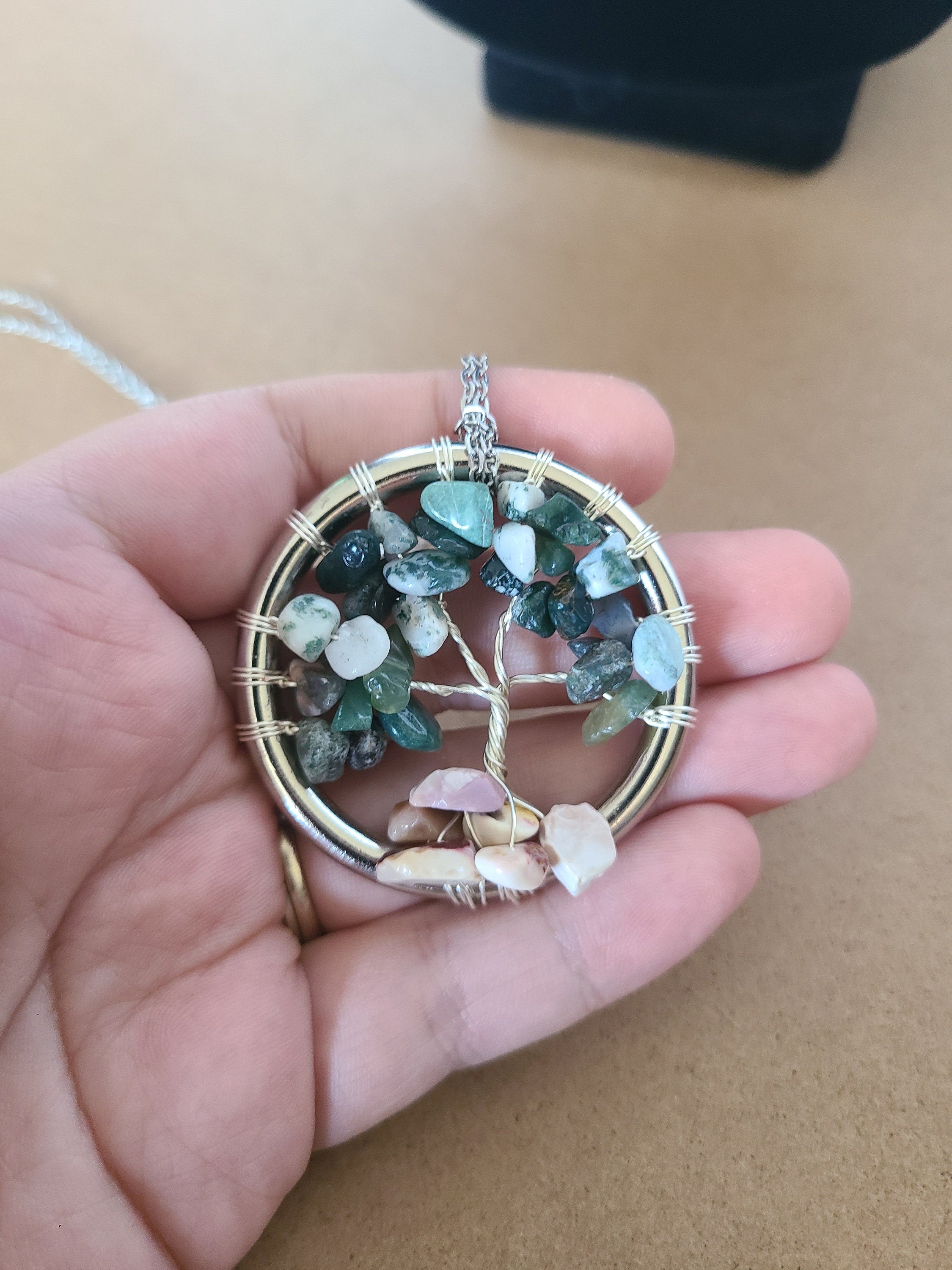 Hand holding a tree of life pendant with multicolored stones against a neutral background
